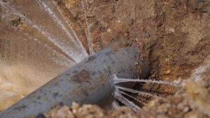 A close-up of a damaged underground pipe with multiple water leaks, showing water spraying out from several cracks in the pipe.